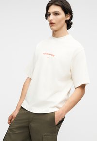 Mladić tamne kose, obučen u bijelu majicu s natpisom "HÔTEL AMOUR" i maslinasto zelene hlače, stoji s jednom rukom u džepu.