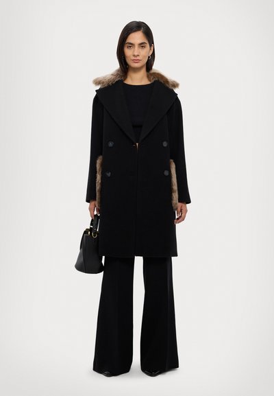 Cappotto nero doppiopetto con ampio colletto e rifiniture in pelliccia sintetica, caratterizzato da sei bottoni, abbinato a pantaloni neri a gamba larga e una borsa nera.