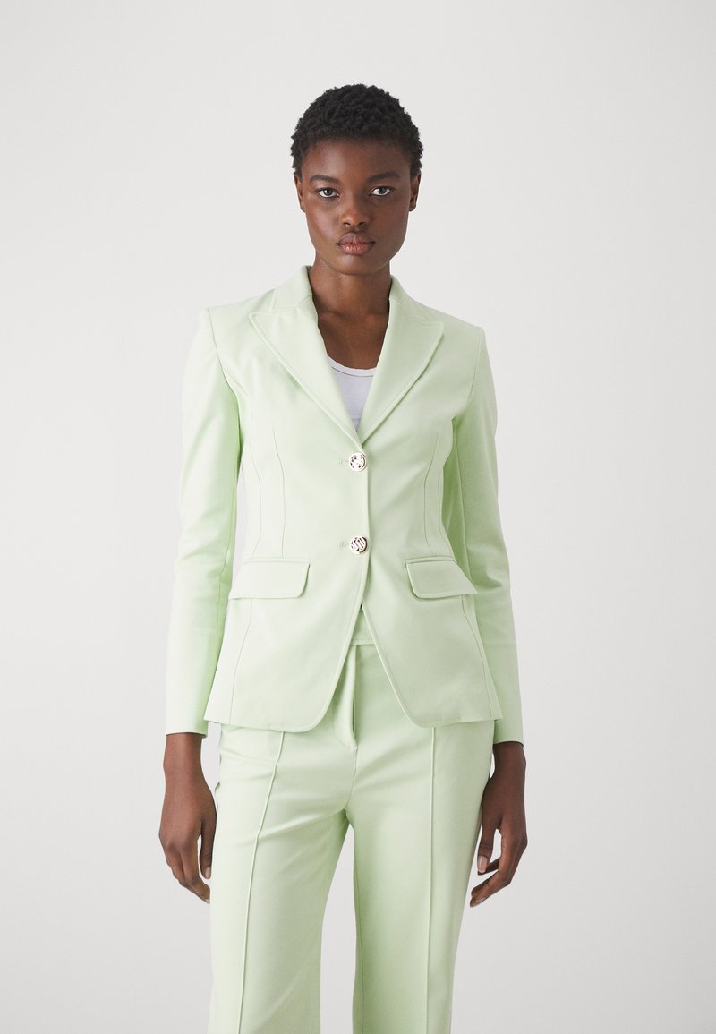 Blazer verde chiaro con design aderente, spalle strutturate, due tasche frontali e due bottoni decorativi, abbinato a pantaloni coordinati.
