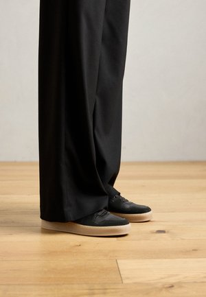 Personne portant un pantalon noir large et des baskets noires avec des semelles épaisses beiges se tenant sur un sol en bois clair.