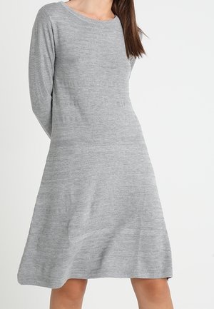 Kobieta ubrana w jasnoszarą dzianinową sukienkę do kolan z długim rękawem, z delikatnymi poziomymi teksturowanymi paskami na białym tle.