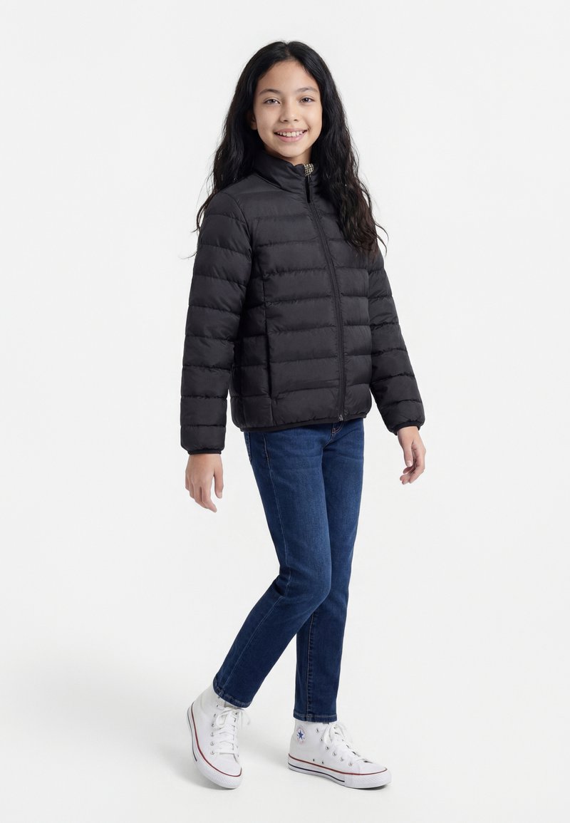 Mädchen mit langem dunklem Haar lächelt, trägt eine schwarze Daunenjacke, blaue Jeans und weiße hohe Sneaker und steht vor einem weißen Hintergrund.