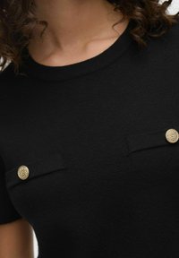 Svart stickad topp med korta ärmar, rund halsringning och två framfickor med guld, runda dekorativa knappar. Slät textur.