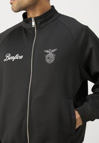 Sort zip-up jakke med høj krave, forsynet med et hvidt broderet "Benfica" logo og emblem, glat stof og sidelommer.