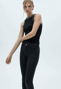 Črna brezrokavnik s belimi detajli preko črnih skinny kavbojk. Top ima visok ovratnik in prilegajočo zasnovo. Gladke teksture skozi celotno obleko.