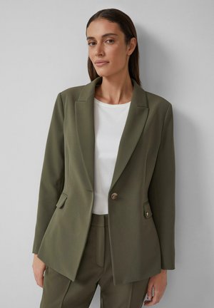 Blazer strutturato verde oliva con un solo bottone, risvolti affilati e due tasche frontali, abbinato a una t-shirt bianca. Tessuto con una texture liscia.