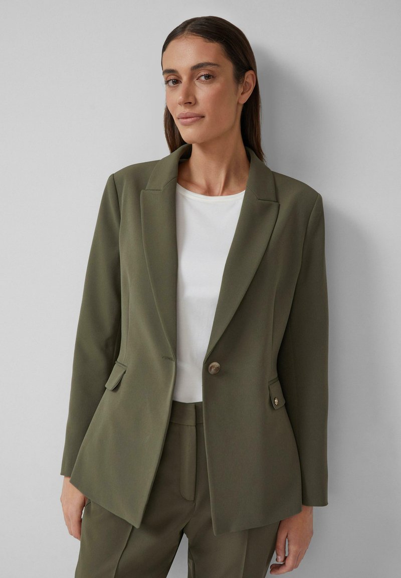 Blazer estructurado en verde oliva con un solo botón, solapas afiladas y dos bolsillos frontales, combinado con una camiseta blanca. Textura de tela suave.