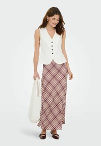 Gilet sans manches blanc avec des boutons noirs, associé à une longue jupe à carreaux dans des tons de bordeaux et crème. Tient un sac fourre-tout blanc texturé.