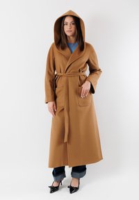Cappotto lungo color marrone cammello, con cappuccio e cintola, aperto sul davanti, con due tasche laterali e maniche taperate, indossato sopra jeans e scarpe con tacchi alti.