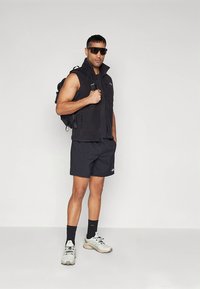 Musta fleeceliivi lyhythihaisen paidan päällä, yhdistetty mustiin shortseihin. Asusteina aurinkolasit ja reppu. Vaaleat urheiluvalkoiset kengät.