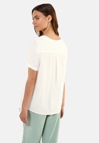 Blusa bianca a maniche corte con scollatura rotonda, caratterizzata da pieghe sul retro e un dettaglio legato sul lato. Tessuto morbido e liscio.