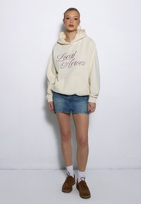 Kremowa bluza z kapturem z napisem "Local Heroes", jeansowa mini spódnica, białe skarpetki i brązowe buty. Miękki materiał, luźny krój i design z kapturem.