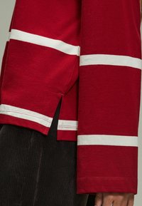 Rotes Langarmshirt mit weißen horizontalen Streifen, ausgestattet mit seitlichen Schlitzen und genähten Saumdetails. Das Material wirkt weich und strukturiert.