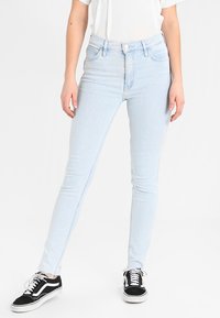 Jeans ajustados de color azul claro hechos de denim con un diseño clásico de cinco bolsillos. Llevados con una camiseta blanca y zapatillas negras.