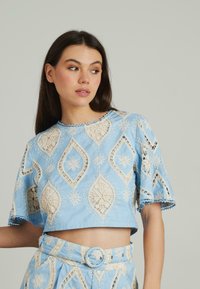 Blaues cropped Top mit aufwendiger cremefarbener Spitzenstickerei in diamant- und blumenförmigen Mustern, mit kurzen Ärmeln und gewellten Kanten.