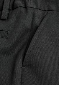 Pantalons en tissu noir avec une texture lisse, présentant une couture détaillée le long de la couture et une ouverture de poche latérale.