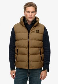 Gilet imbottito marrone con chiusura a zip e cappuccio, caratterizzato da una patch con logo e due tasche laterali, indossato sopra un maglione a trecce blu navy.