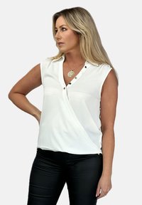 Biała bezrękawnikowa bluzka z fasonem na zakładkę, z przodu z guzikami i subtelnym połyskiem. Połączona z czarnymi dopasowanymi spodniami. Gładki materiał o minimalnej teksturze.