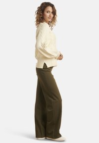 Cream cable-knit sweater with ribbed texture, side slits; paired with olive wide-leg trousers and white sneakers, seen from the side.