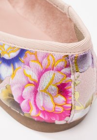 Chaussure plate florale présentant une toile rose avec des motifs de fleurs multicolores, une finition texturée et une doublure intérieure douce. Semelle marron.