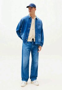 Jovem vestido com uma jaqueta de denim azul e jeans, camisa bege com botões, boné azul e sapatilhas brancas, de pé com as mãos atrás das costas.
