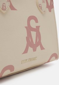 Sac à main beige avec lettres SM roses entrelacées et logo Steve Madden doré sur une surface en cuir texturé.