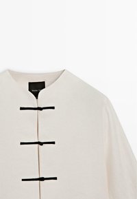 Hvid beklædning med rund krave og tre sorte knudefrogbeslag ned langs midten foran.