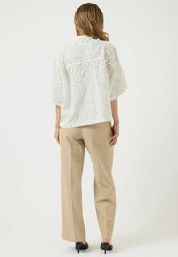 Blusa bianca ricamata con maniche corte a palloncino, caratterizzata da motivi a intaglio, abbinata a pantaloni beige su misura e tacchi neri.