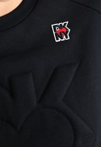 Felpa nera con un design del logo in rilievo e testurizzato. Presenta un piccolo logo ricamato in bianco, rosso e nero sul petto.