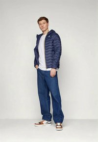 Junger Mann, der eine marineblaue gesteppte Jacke, ein weißes T-Shirt, lockere blaue Jeans und braun-weiße Sneakers trägt, vor einem schlichten hellen Hintergrund steht.