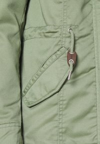 Primo piano della tasca di una giacca verde oliva con patta e cordino, dotata di un aglet in metallo e di un fermaglio in pelle.