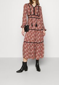 Maxikleid im roten Blumenmuster mit schwarzen Akzenten, langen Ärmeln und V-Ausschnitt. Mit Bindedetails und einem gestuften Design, kombiniert mit schwarzen Stiefeln.