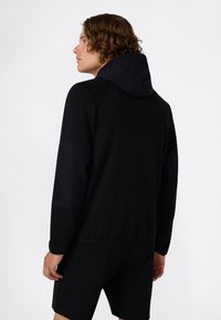 Hombre con cabello castaño ondulado que lleva una sudadera negra con capucha, combinada con pantalones cortos negros, de pie contra un fondo blanco liso.