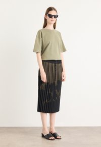 T-shirt oversize in verde oliva in cotone abbinato a una gonna plissettata nera con texture in rete e dettagli dorati; mules nere con punta aperta.