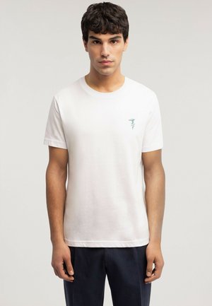 Joven con cabello rizado oscuro que lleva una camiseta blanca de manga corta con un pequeño logo azul en el pecho izquierdo, de pie frente a un fondo liso.