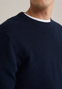 Mörkblå texturerad stickad tröja med rund hals över ett vitt innerlager. Synliga sömmar och ribbade muddar. Närbild.