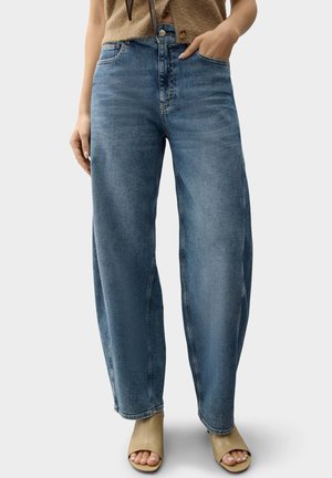 Locker sitzende, blaue Jeans mit niedrigem Bund, getragen mit beigen offenen Absätzen und einem braunen ärmellosen Oberteil, Hand in der Tasche, vor einfachem Hintergrund.