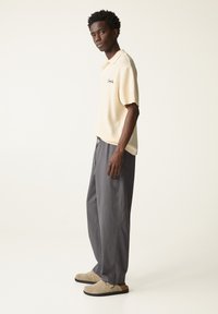 Cremfarbener Polo-Shirt mit Struktur und kurzen Ärmeln, kombiniert mit grauen Jogginghosen mit Kordelzug und beigen Slippern. Schlichtes Design, entspannte Passform.