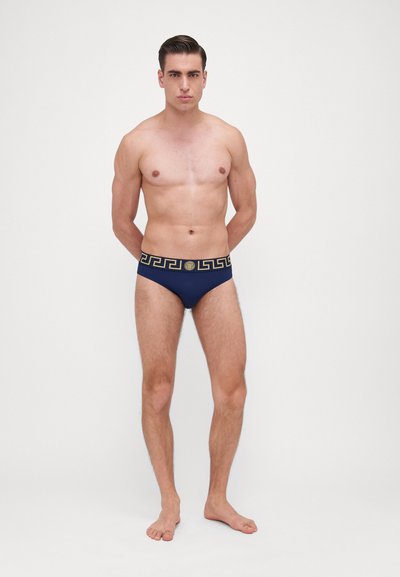 Marine svømmebukser med et gullfarget gresk nøkkelmønster på den elastiske midjebåndet. Stoffet ser glatt og fleksibelt ut. Modellen står barføtt.