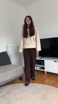 Beżowa bluza polarowa z kapturem i kieszeniami, połączona z luźnymi brązowymi spodniami i czarnymi butami, na jasnym dywanie w salonie.