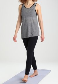 Grijs geribd tanktop met zwarte accenten en een klein logo, gecombineerd met zwarte leggings. Het model staat op blote voeten op een lichtgekleurde yogamat.