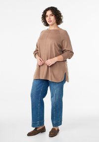 Béžový pletený sveter s okrúhlym výstrihom a bočnými rozparkami, doplnený svetlomodrými džínsami a hnedými mokasínami.