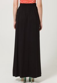 Jupe maxi noire avec une taille élastique, texture lisse, design fluide et coupe longueur cheville, associée à des talons turquoise.