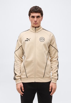 Vyras dėvintis smėlio spalvos Puma striukę su užtrauktuku, su BVB 09 logotipu ir užrašu „KING“, stovintis prieš paprastą šviesią foną.