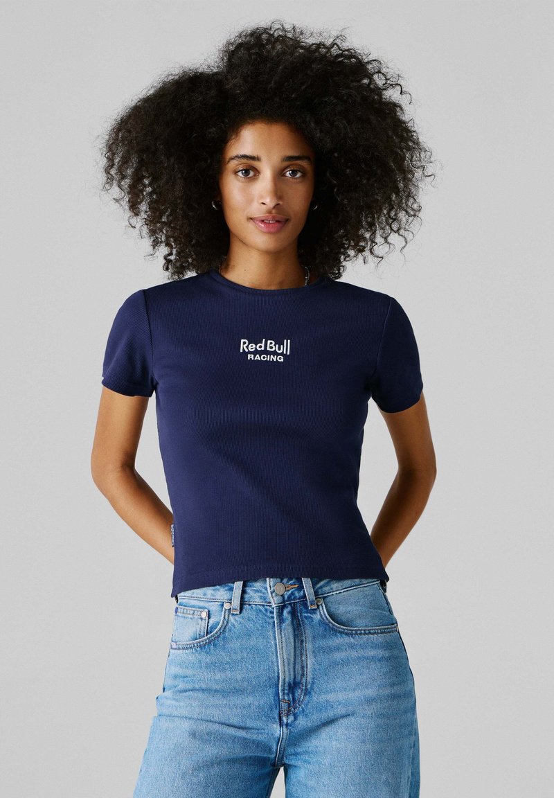 Γυναίκα με σγουρά μαλλιά που φοράει μπλε-σκούρο μπλουζάκι Red Bull Racing και ανοιχτόμπλε τζιν, στέκεται με τα χέρια πίσω από την πλάτη της.