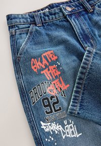 Jean en denim bleu avec texte imprimé "SKATE THE CITY" en rouge, "BROOKLYN 92 NEW YORK" en noir et des accents style graffiti blancs sur la cuisse.