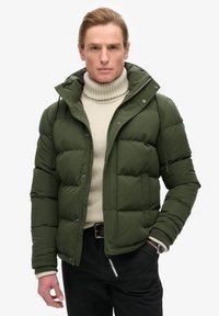 Giacca a sacco verde oliva con cappuccio rimovibile, chiusura con zip e bottoni, e polsini a coste, indossata sopra un dolcevita crema. Pantaloni neri.