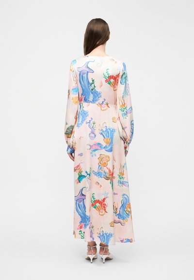 Γυναίκα με μακριά καστανά μαλλιά, φορεμένη με μια μακριά ροζ παστέλ φόρεμα με πολύχρωμα σχέδια θαλάσσιων ζώων, που στέκεται μπροστά σε μια απλή γκρι φόντο.