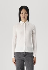 Long-sleeve white shirt with a collar, ribbed texture, and button closure, paired with dark gray pants, standing against a plain background.