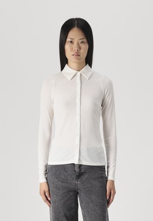 Wit overhemd met lange mouwen en een kraag, geribbelde textuur en knoopsluiting, gecombineerd met donkergrijze broek, staand tegen een eenvoudige achtergrond.